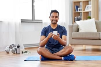 sport, technology and healthy lifestyle concept - smiling indian man with smartphone sitting on exercise mat at home. indian man with smartphone on exercise mat at home