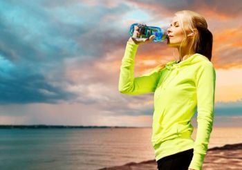 fitness, sport and healthy lifestyle concept - woman with earphones drinking water after exercising over sea sunset background. woman drinking water after exercising at seaside
