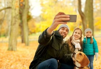family, pets and people concept - happy mother, father and little daughter with beagle dog taking selfie by smartphone over autumn park background. happy family with dog taking selfie in autumn park
