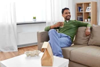 consumption and people concept - smiling indian man with takeaway coffee and food at home. indian man with takeaway coffee and food at home