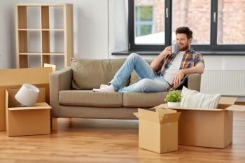 moving, people and real estate concept - happy man with boxes sitting on sofa and drinking coffee at new home. man with boxes and drinking coffee at new home