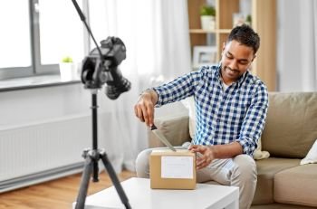 mail delivery, shipment and blogging concept - happy indian male blogger opening parcel box with knife and recording unboxing video with camera at home. male video blogger opening parcel box at home