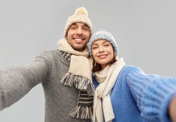 people, christmas and winter clothes concept - happy couple in knitted hats and scarves taking selfie over grey background. happy couple in winter clothes taking selfie