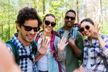 travel, tourism, hike and people concept - group of friends with backpacks taking selfie and waving hands in forest. friends with backpacks hiking and taking selfie