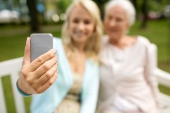 family, technology and people concept - close up of happy smiling adult daughter and senior mother with smartphone sitting on park bench and taking selfie. daughter and senior mother taking selfie at park