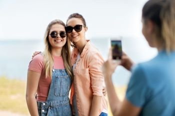 leisure and friendship concept - happy smiling teenage girls or best friends in sunglasses being photographed by smartphone at seaside in summer. teenage girls or best friends being photographed