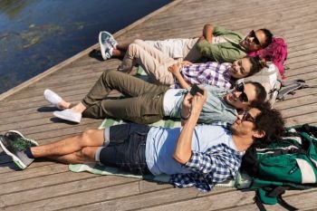 friendship, technology and travel concept - group of friends friends with smartphone and backpacks lying on lake pier. friends with smartphone lying on lake pier