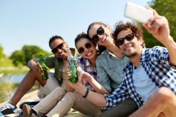 leisure, picnic and technology concept - friends with drinks taking selfie by smartphone on lake pier in summer park. friends with drinks taking selfie by smartphone
