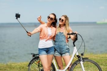 leisure, technology and friendship concept - happy smiling teenage girls or friends with bicycle taking picture by selfie stick on smartphone at seaside in summer. teenage girls with bicycle taking selfie in summer