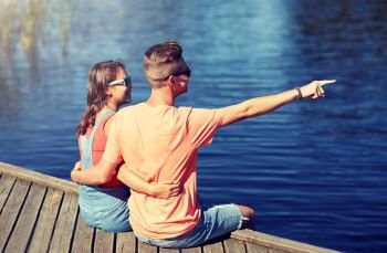 holidays, vacation, love and people concept - happy smiling teenage couple hugging and pointing finger to something on river berth at summer. happy couple pointing finger on summer river berth