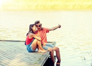 holidays, vacation, love and people concept - happy teenage couple taking selfie on smartphone and sitting on river berth at summer. happy teenage couple taking selfie on smartphone