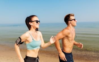 fitness, sport and technology concept - happy couple with earphones and arm bands running along summer beach. couple with phones and arm bands running on beach