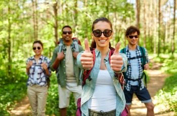 travel, tourism, hike and people concept - group of friends with backpacks and woman showing thumbs up gesture in forest. friends with backpacks showing thumbs up in forest