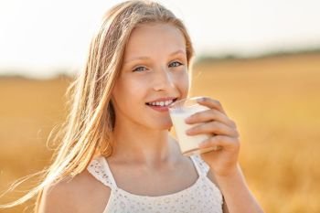 nature, healthy eating and organic concept - smiling young girl drinking milk from glass on cereal field in summer. girl drinking milk from glass on cereal field