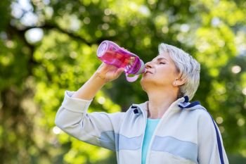 fitness, sport and healthy lifestyle concept - thirsty senior woman drinking water from bottle after exercising in park. senior woman drinks water after exercising in park