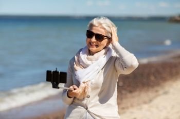 old people and leisure concept - happy smiling senior woman taking picture by smartphone and selfie stick on beach in estonia. senior woman taking selfie on beach