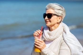 people and leisure concept - senior woman in sunglasses drinking shake, orange juice or smoothie on beach in estonia. senior woman drinking orange juice on beach