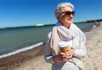 people and leisure concept - senior woman in sunglasses drinking takeaway coffee on beach in estonia. senior woman drinking takeaway coffee on beach