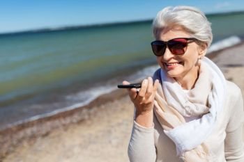 old people and leisure concept - happy smiling senior woman using voice command recorder on smartphone, on beach in estonia. old woman recording voice by smartphone on beach