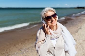 technology, old people and leisure concept - senior woman in headphones and sunglasses listening to music on summer beach in estonia. old woman in headphones listens to music on beach