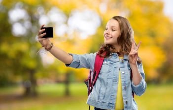 education, school and people concept - happy smiling teenage student girl with bag taking selfie by smartphone and showing peace over autumn park background. teenage student girl taking selfie by smartphone
