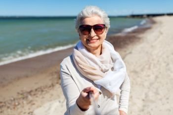 old people and leisure concept - happy smiling senior woman in sunglasses taking picture by selfie stick on beach in estonia. old woman taking picture by selfie stick on beach