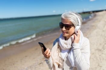 technology, old people and leisure concept - senior woman in headphones and sunglasses listening to music on smartphone on summer beach in estonia. old woman in headphones with smartphone on beach