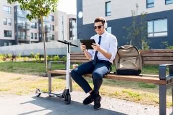 business, technology and people concept - young smiling businessman with tablet computer, headphones, backpack and electric scooter sitting on street bench in city. businessman with tablet pc, headphones and scooter