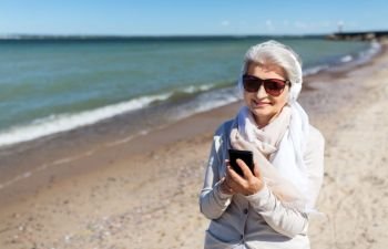 technology, old people and leisure concept - senior woman in headphones and sunglasses listening to music on smartphone on summer beach in estonia. old woman in headphones with smartphone on beach
