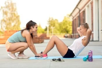 fitness, sport and healthy lifestyle concept - young woman assisting her friend doing sit-ups on mat outdoors. women training and doing sit-ups outdoors