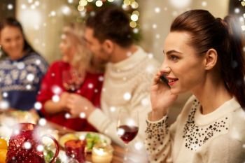 holidays, communication and celebration concept - close up of young woman calling on smartphone and having christmas dinner with friends at home over snow. woman calling on smartphone at christmas dinner