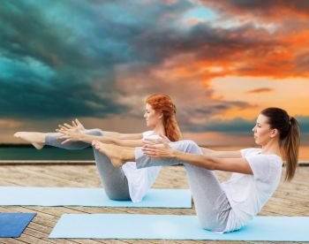 fitness, sport, yoga, people and healthy lifestyle concept - women making half-boat pose on mat outdoors on sea pier over sunset background. women making yoga in half-boat pose outdoors