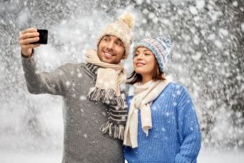 people, christmas and winter clothes concept - happy couple in knitted hats and scarves taking selfie by smartphone over snow background. couple in winter hats taking selfie by smartphone