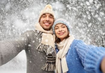 people, christmas and winter clothes concept - happy couple in knitted hats and scarves taking selfie over snow background. happy couple in winter clothes taking selfie