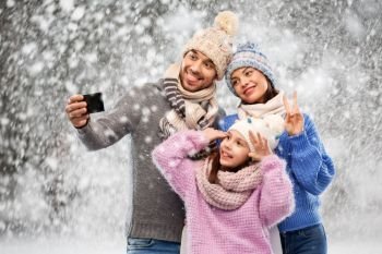 family, christmas and technology concept - happy mother, father and little daughter in winter clothes taking selfie by smartphone over snow background. happy family taking selfie by smartphone over snow