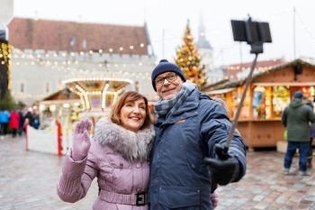 technology, winter holidays and people concept - happy senior couple taking picture by smartphone on selfie stick at christmas market on town hall square in tallinn, estonia. senior couple taking selfie at christmas market
