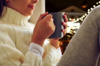 hot drinks, holidays and people concept - close up of beautiful girl in winter sweater with tea mug sitting at window over christmas lights background. close up of girl with tea mug sitting at window