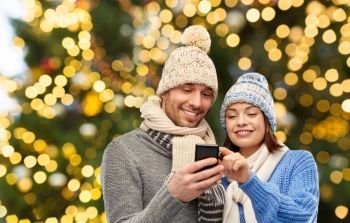 technology, christmas and winter clothes concept - happy couple in knitted hats and scarves with smartphone over festive lights background. happy couple with smartphone over christmas lights