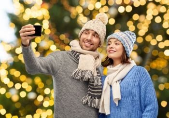 people, christmas and winter clothes concept - happy couple in knitted hats and scarves taking selfie by smartphone over festive lights background. couple taking selfie by smartphone on christmas