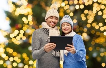 technology, christmas and winter clothes concept - happy couple in knitted hats and scarves with tablet computer over festive lights background. couple with tablet computer over christmas lights