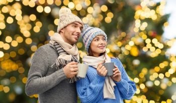 people, christmas and love concept - happy romantic couple in knitted hats and scarves with mugs over festive lights background. happy couple with mugs over christmas lights