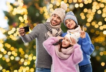 family, christmas and technology concept - happy mother, father and little daughter in winter clothes taking selfie by smartphone over festive lights background. happy family taking selfie over christmas lights