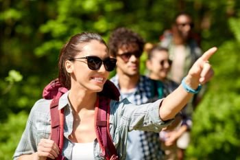 travel, tourism, hike and people concept - woman showing something to group of friends walking with backpacks in forest. group of friends with backpacks hiking in forest