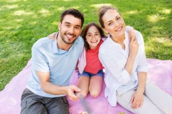 family, leisure and technology concept - happy mother, father and daughter having picnic and taking picture by selfie stick at summer park. family having picnic and taking selfie at park
