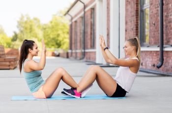 fitness, sport and healthy lifestyle concept - smiling young women or female friends doing sit-ups and making high five outdoors. happy women doing sit-ups and making high five