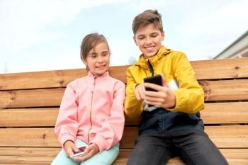 childhood, technology and people concept - happy children or brother and sister smartphones sitting on wooden street bench outdoors. children with smartphones sitting on street bench