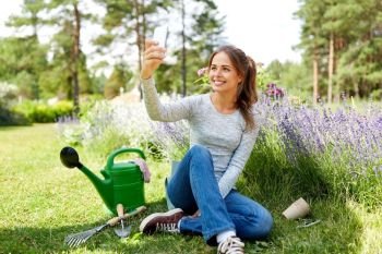 gardening, technology and people concept - happy young woman or gardener with garden tools taking selfie by smartphone in summer. woman taking selfie by smartphone in summer garden