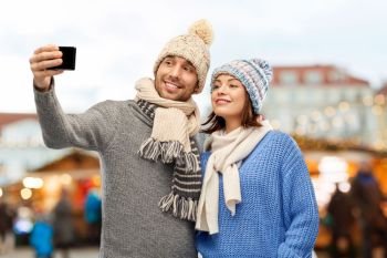 people, technology and winter holidays concept - happy couple in knitted hats and scarves taking selfie by smartphone over christmas market in old town of tallinn city background. couple takes selfie by smartphone christmas market