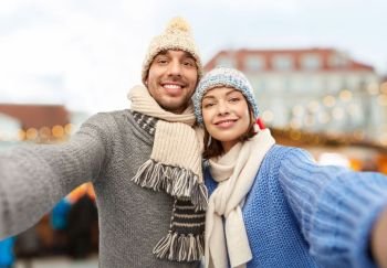 people and winter holidays concept - happy couple in knitted hats and scarves taking selfie over christmas market in old town of tallinn city background. happy couple taking selfie at christmas market