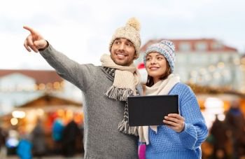 technology and winter holidays concept - happy couple in knitted hats and scarves with tablet computer over christmas market in old town of tallinn city background. couple with tablet computer at christmas market